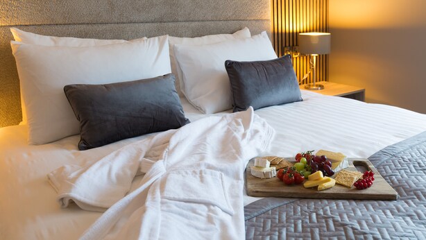 A neatly made bed with white and gray bedding, a wooden nightstand with a lamp, and a fruit and cheese board on the bed. - Castle Varagh Hotel & Bar (Castlepollard)