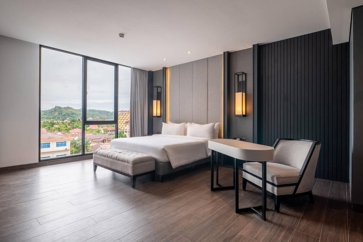 Une chambre d’hôtel moderne avec un grand lit, une petite table et une chaise. De grandes fenêtres offrent une vue sur les bâtiments environnants.