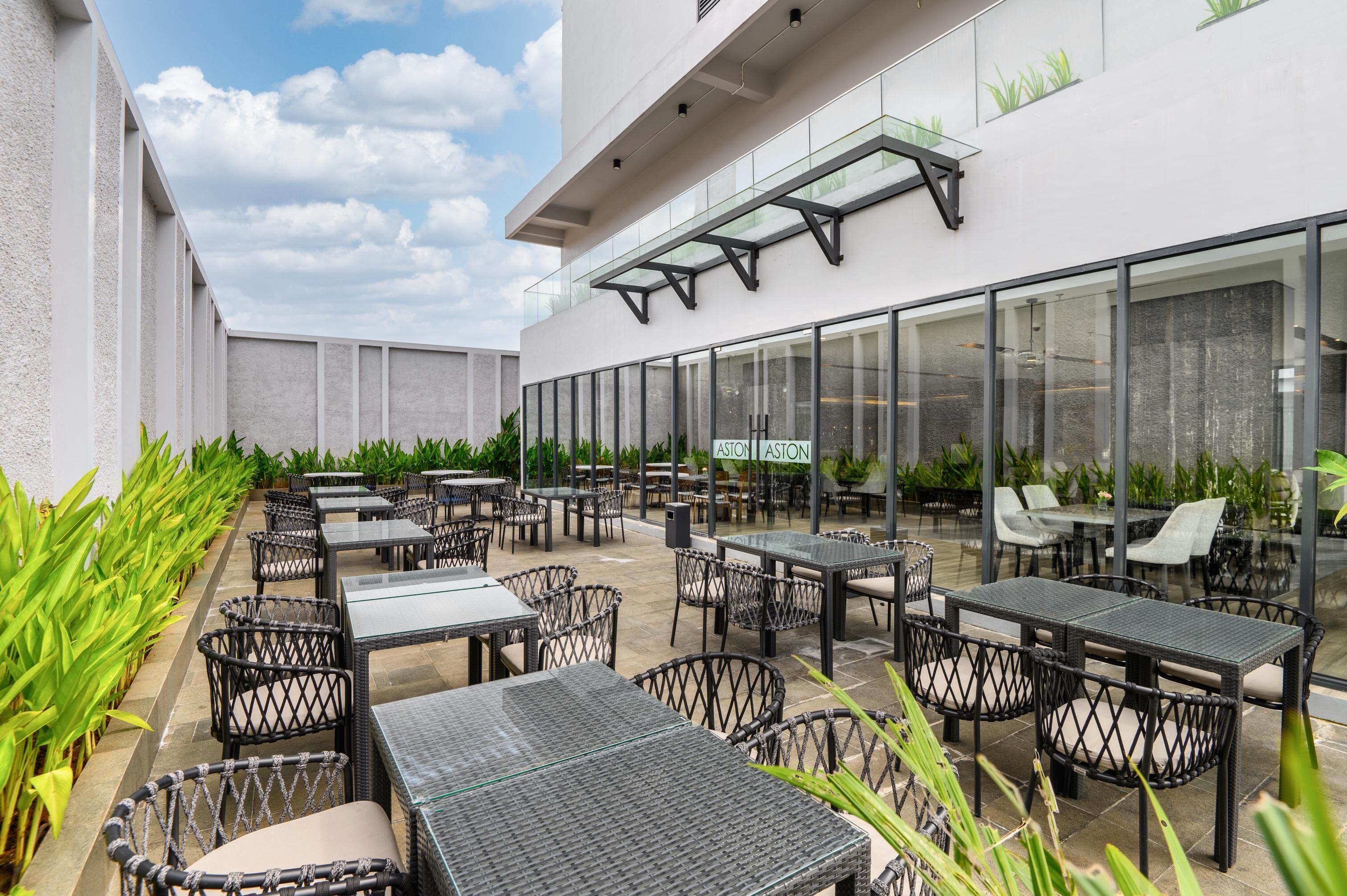 Un espace extérieur aménagé en terrasse, avec plusieurs tables et chaises, entouré de murs de verre et de plantes.