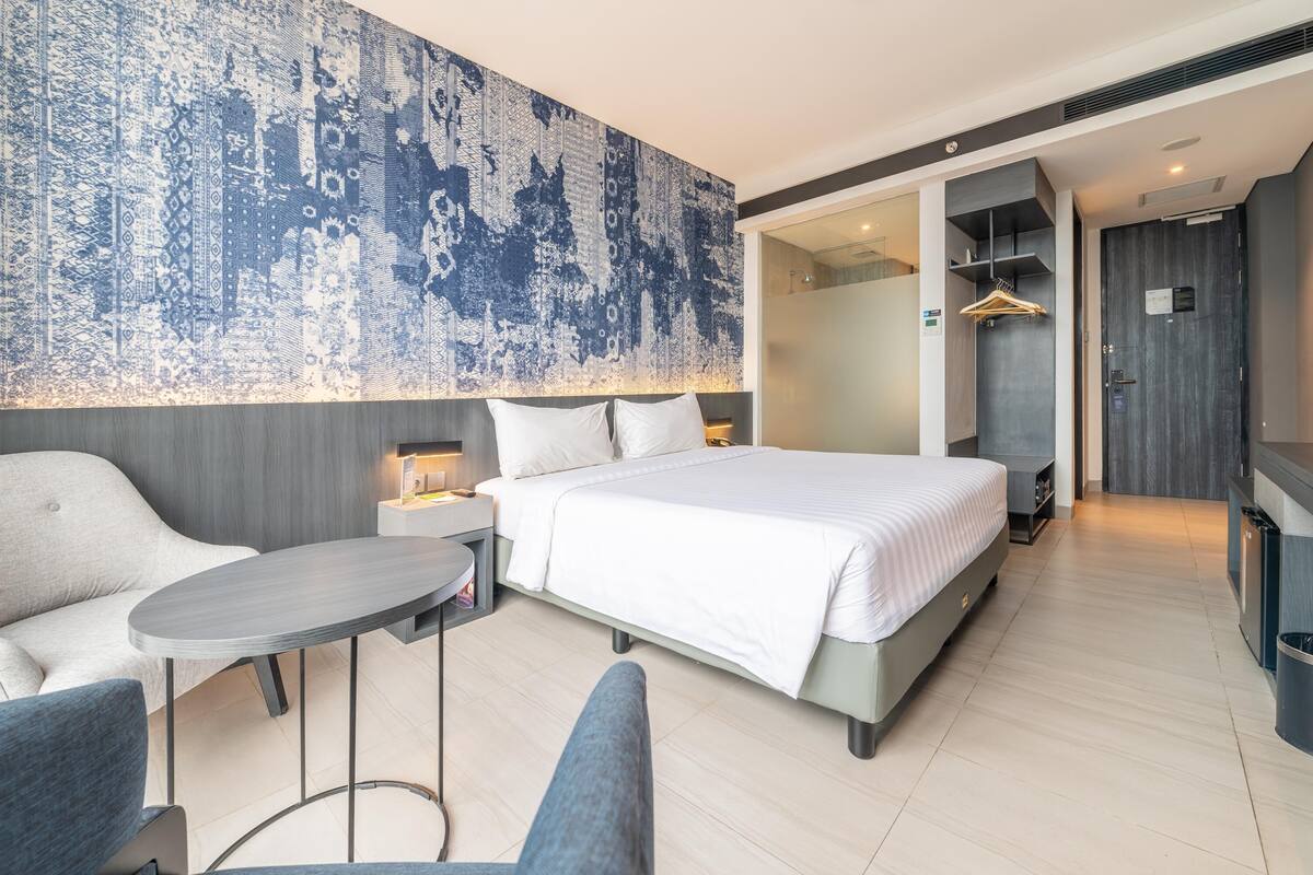 Une chambre d’hôtel moderne avec un grand lit, une petite table et un canapé. Un grand tableau abstrait orne le mur, et une armoire avec un miroir est également présente.