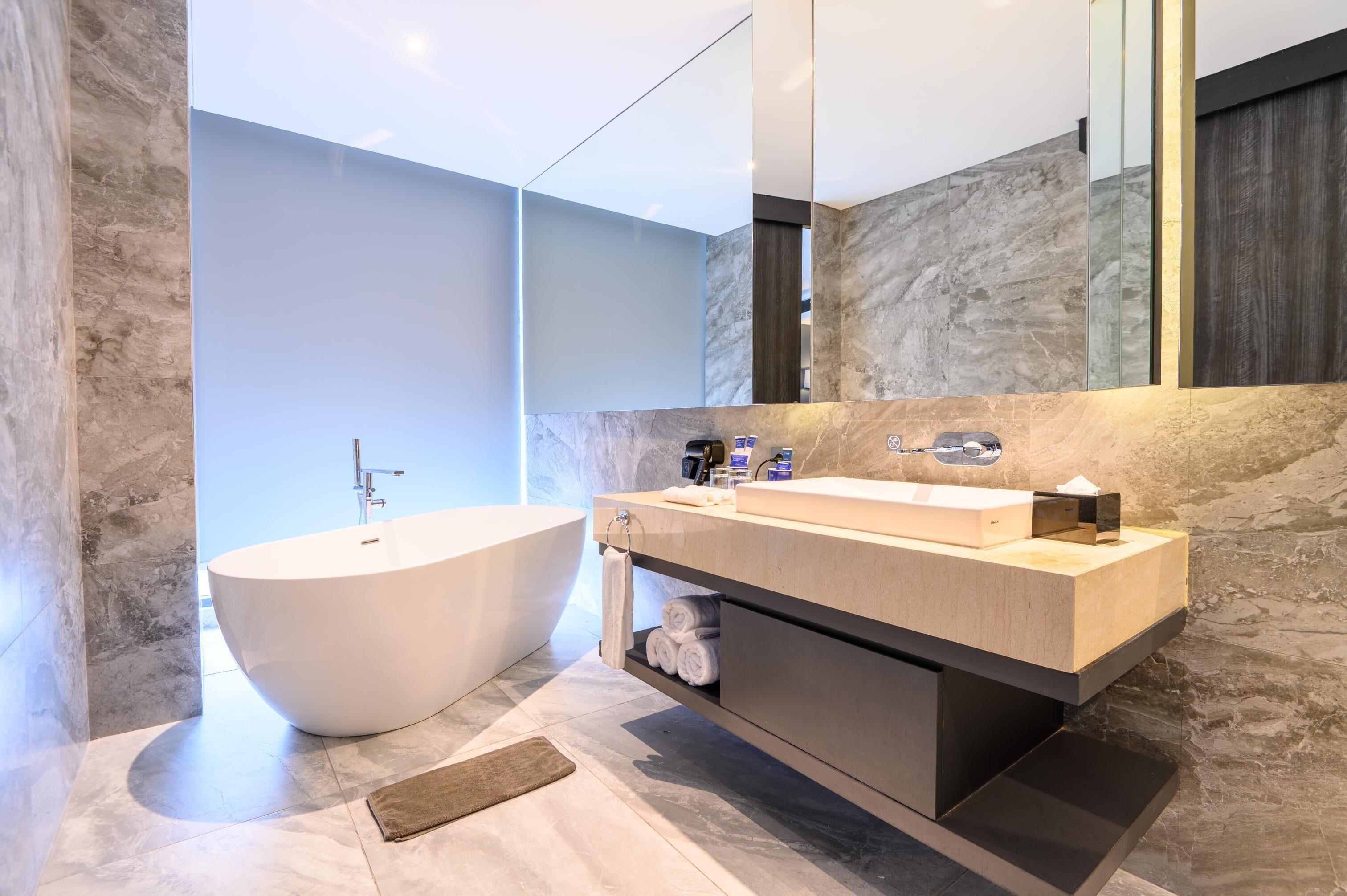 A modern bathroom with a freestanding bathtub, a vanity with a mirror, and marble walls.
