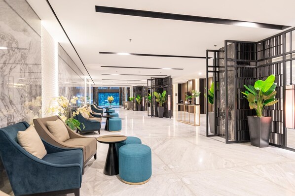 A modern lobby with marble flooring, blue seating, potted plants, and a large TV screen.