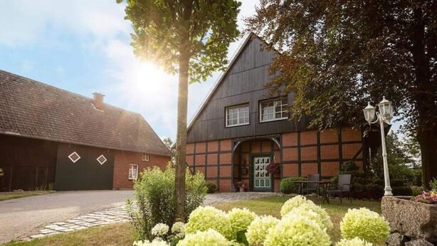 A traditional half-timbered house with a stone foundation, a visible garage, and a well-maintained garden with hydrangeas. - In Idyllischer Wohnlage auf Einem Fachwerkhof in der Natur (Ladbergen)
