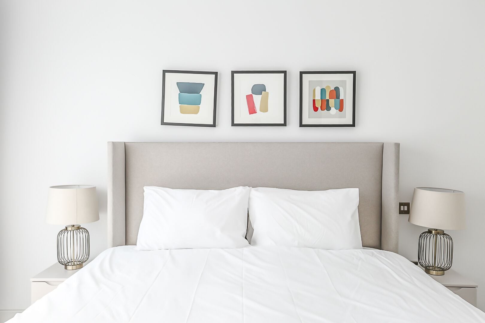 Uma cama bem arrumada com lençóis brancos, duas luminárias de cabeceira e três quadros na parede.