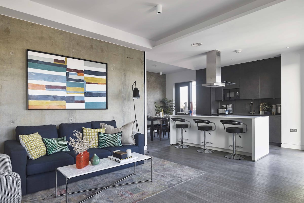 A modern living room with a dark blue sofa, patterned cushions, and a coffee table with decorative items. A large abstract painting adorns the wall, and a kitchen area with dark cabinetry and a bar stool is visible in the background.