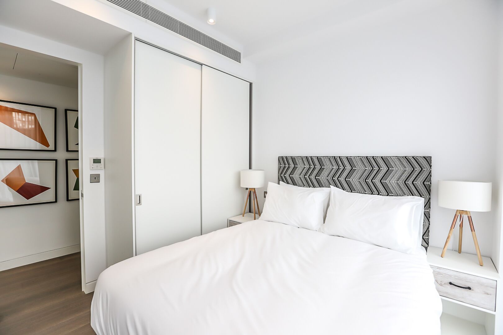 A modern hotel room with a large bed, two bedside tables with lamps, a wardrobe, and abstract wall art.