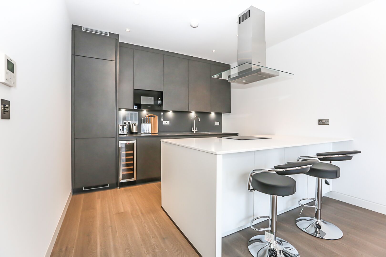 Una cucina moderna con mobili scuri, una cappa aspirante in acciaio inossidabile e un piano di lavoro bianco. Sono presenti due sgabelli da bar con seduta nera e gambe cromate.