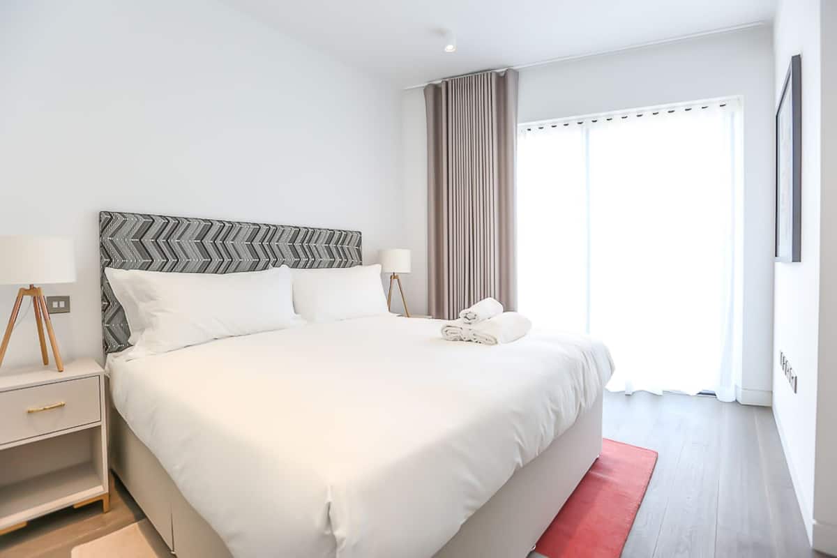A modern hotel room with a large bed, a bedside table with a lamp, and a view of the outside through a window with curtains.