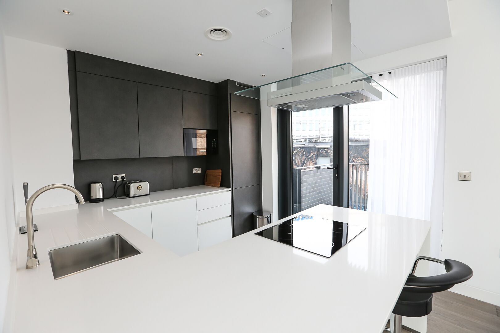A modern kitchen with a white countertop, dark cabinets, a built-in oven, and a glass door leading to a balcony.
