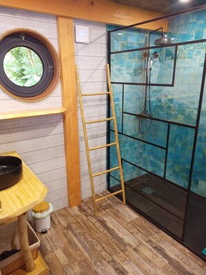 Bathroom features a shower area with a glass door and elevated showerhead, a wooden shelf, and a round window.