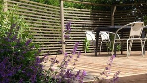 A wooden deck with a pergola, surrounded by lush greenery and purple flowers.