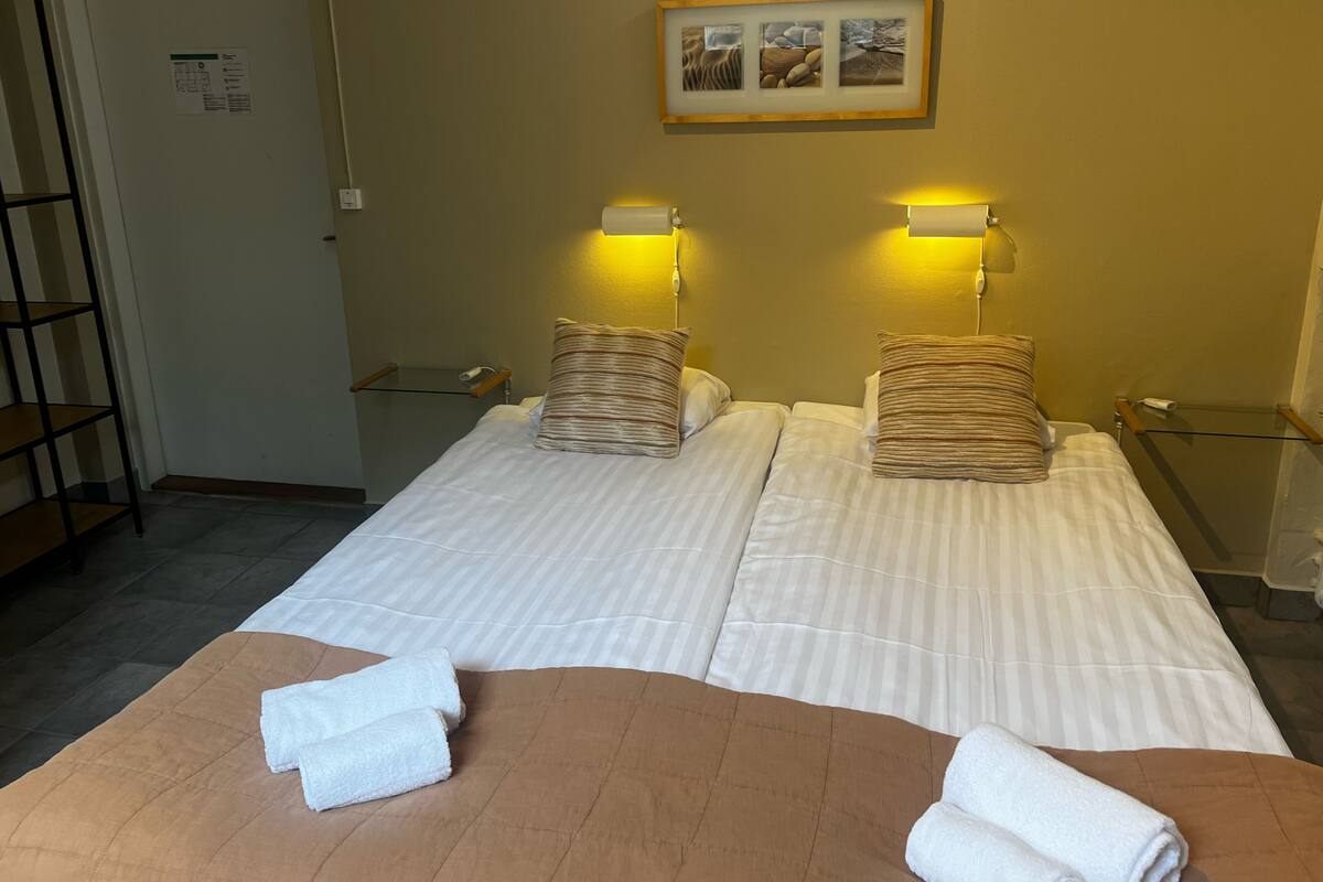 A hotel room with two beds, each with striped bedding and a wooden headboard. There are two bedside lamps, a framed picture on the wall, and a small shelf with a lamp.