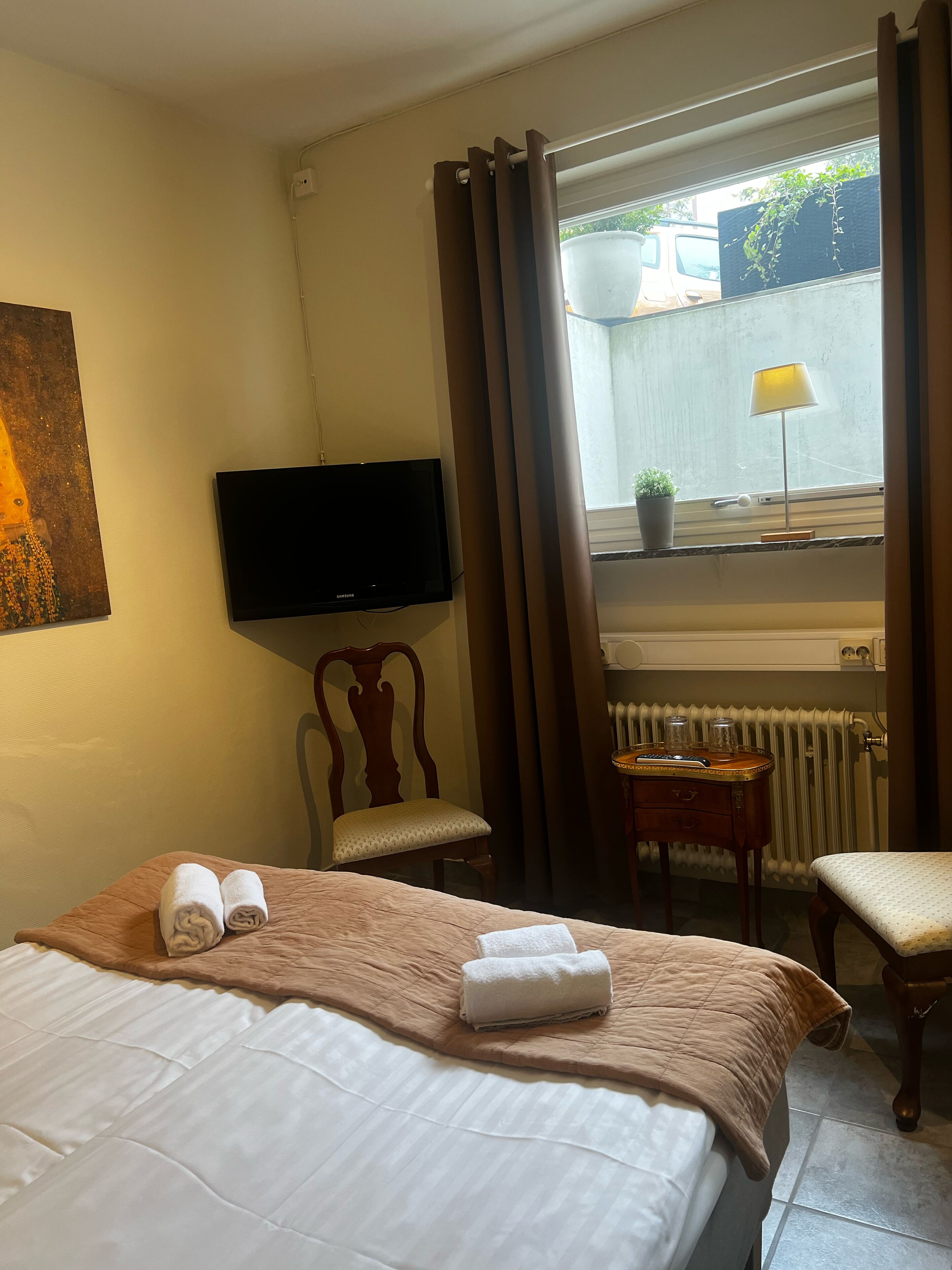 Habitación de hotel con cama, televisor, silla, una mesita de noche con lámpara y ventana con cortinas.