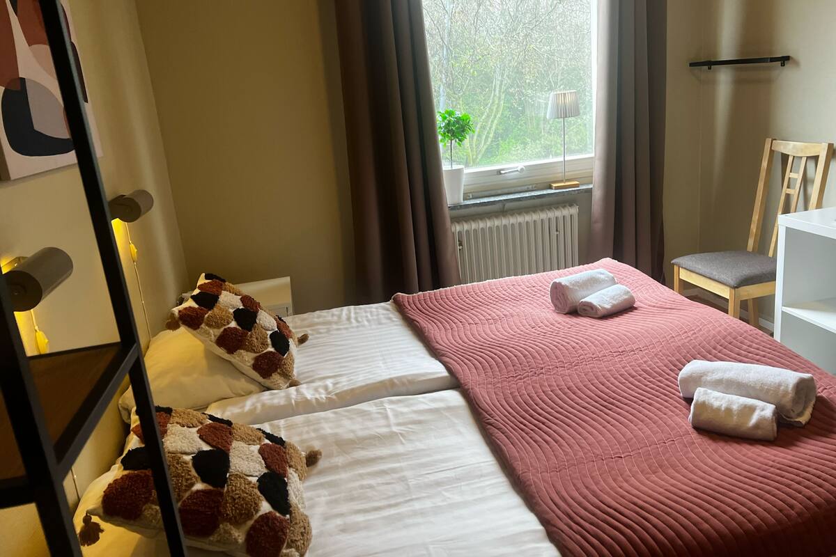A neatly made bed with a pink quilt and two rolled towels, a wooden chair, a white shelf, and a window with a view of trees.