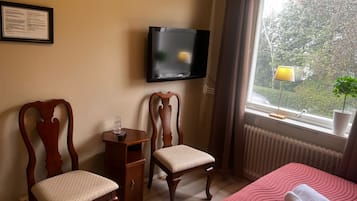 Ein Hotelzimmer mit einem Bett, zwei Holzstühlen, einem kleinen Tisch mit einem Glas und einer Fernbedienung, einem an der Wand montierten Fernseher, einem Fenster mit Blick auf Bäume und einem Heizkörper.