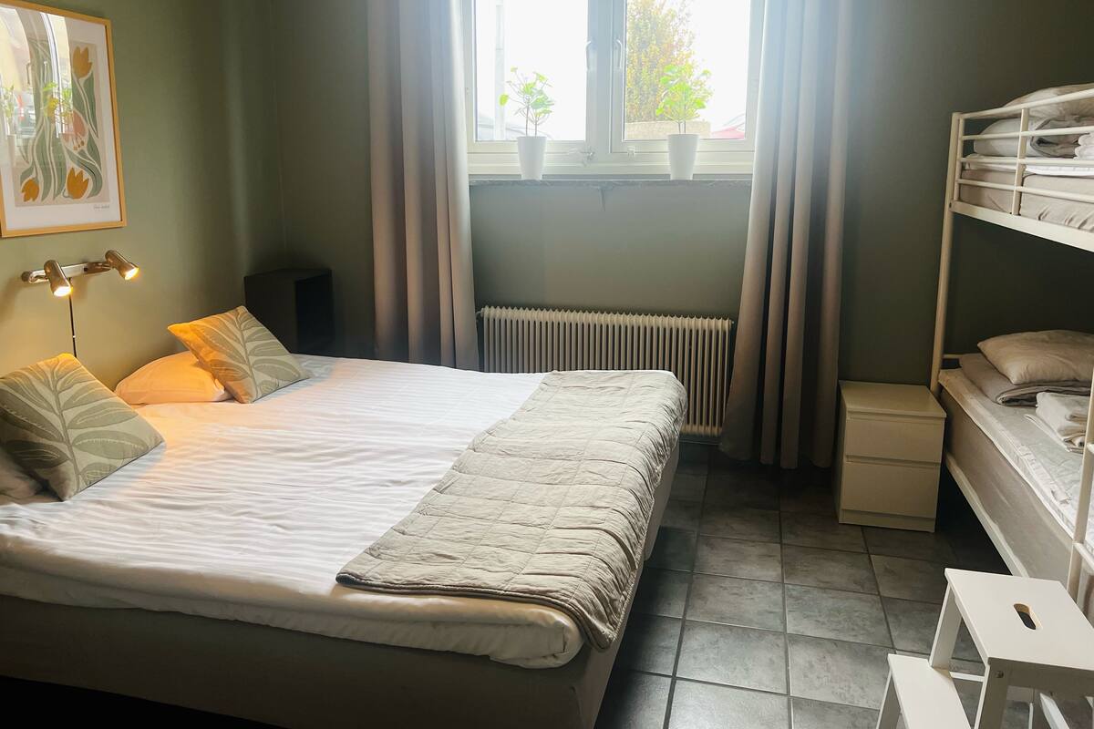 A bedroom with a bed, bedside tables, a radiator, a window with curtains, and a bunk bed.