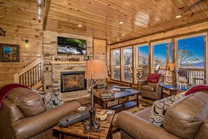 Relax in a warm living room with a stone fireplace, comfortable sofas, and a TV above, offering a view of the outdoors.
