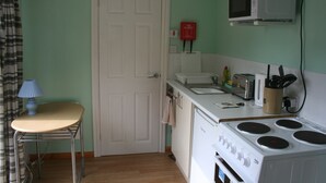 A small kitchen with a microwave, stove, and toaster. There is a door, a chair, and a table. - The Pines Cabin (Swansea)