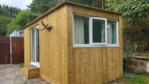 A wooden cabin with a large window and a door, surrounded by a wooden fence and greenery. - The Pines Cabin (Swansea)