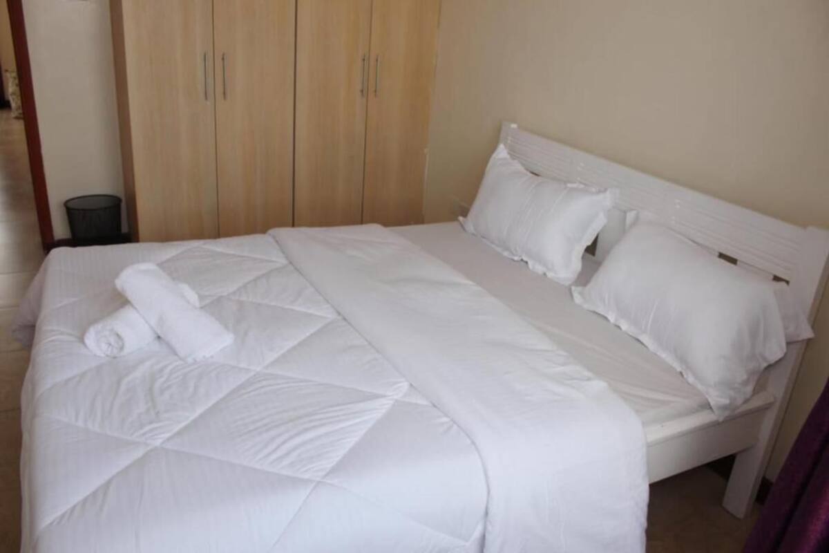 Una cama bien hecha con sábanas y almohadas blancas, un armario de madera y un pequeño cubo de basura.