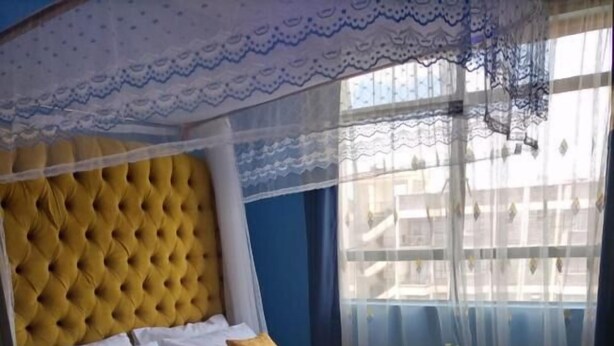 A hotel room with a tufted headboard, a bed with yellow bedding, a bedside table with a lamp, and a window with sheer curtains. - Cosy homes ltd (Mlolongo)