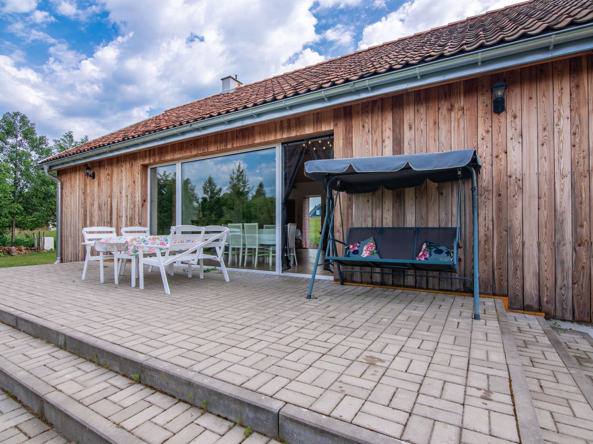 A wooden building with a tiled roof, a covered patio with white chairs and a table, and a swing seat.