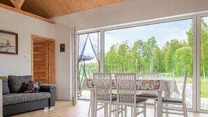 A room with a wooden ceiling, a gray sofa with floral cushions, a dining table with white chairs, and large windows offering a view of trees. - Zagroda nad Krutynią by Interhome (Spychowo-Kierwik)