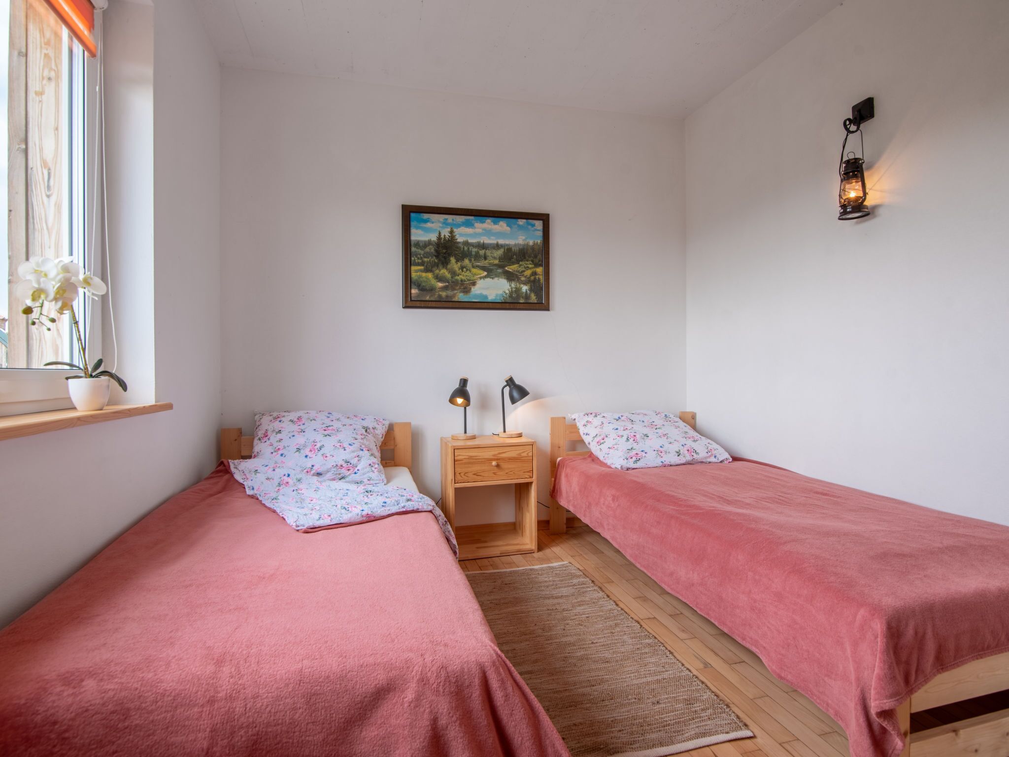 Un dormitorio con dos camas individuales, una mesita de noche con lámparas, una ventana con una planta de orquídea y un cuadro de un paisaje.