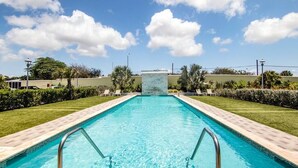 Una piscina rectangular con borde de azulejos, rodeada de césped cuidado y palmeras.