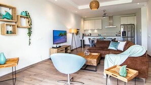 Una sala de estar moderna con un sofá, una mesa de centro y un televisor. Se observa una cocina al fondo con armarios y un refrigerador.