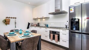 Una cocina moderna con armarios blancos, un refrigerador de acero inoxidable y un comedor con mesa y sillas de madera.