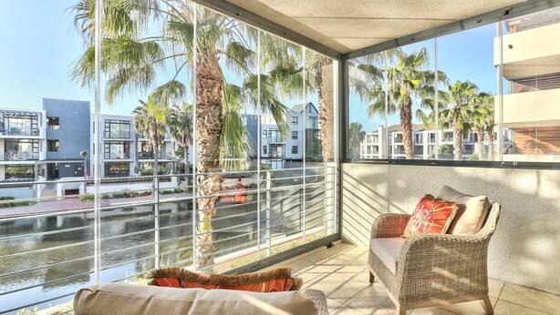 A balcony with wicker furniture, a view of palm trees, and modern buildings in the background. - 2 Bedroom Apartment in Century City (Cape Town)
