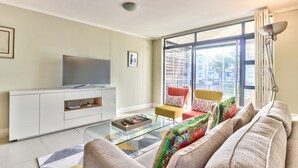 A modern living room with a glass coffee table, a sofa, a television on a white cabinet, and a large window offering a view of buildings. - 2 Bedroom Apartment in Century City (Cape Town)