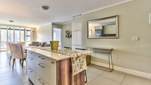 A modern kitchen with a central island, a dining area with chairs, a large mirror, and a decorative wall piece. - 2 Bedroom Apartment in Century City (Cape Town)