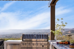 Ein Edelstahlgrill mit mehreren Brennern und einer Steuerung, vor einer malerischen Kulisse aus Hügeln und blauem Himmel.