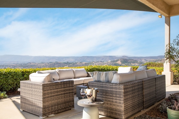 A covered outdoor seating area with wicker furniture and a table, overlooking a scenic landscape. - Architectural Estate w Pool, Spa, Game Room & Sweeping Valley Views | Camila (Temecula)