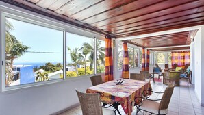 Et overdækket spiseområde med bord og stole, store vinduer og udsigt til havet.