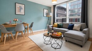 A modern living room with a grey sofa, a dining table with chairs, a coffee table with fruit, and a large window with a city view.