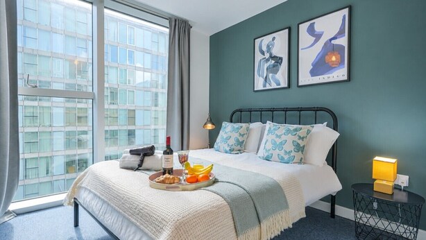 A hotel room with a bed, a bedside table with a lamp, a tray with food and drink, and large windows with a city view.