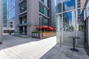 A modern building with large windows, outdoor seating with red umbrellas, and a glass door entrance.