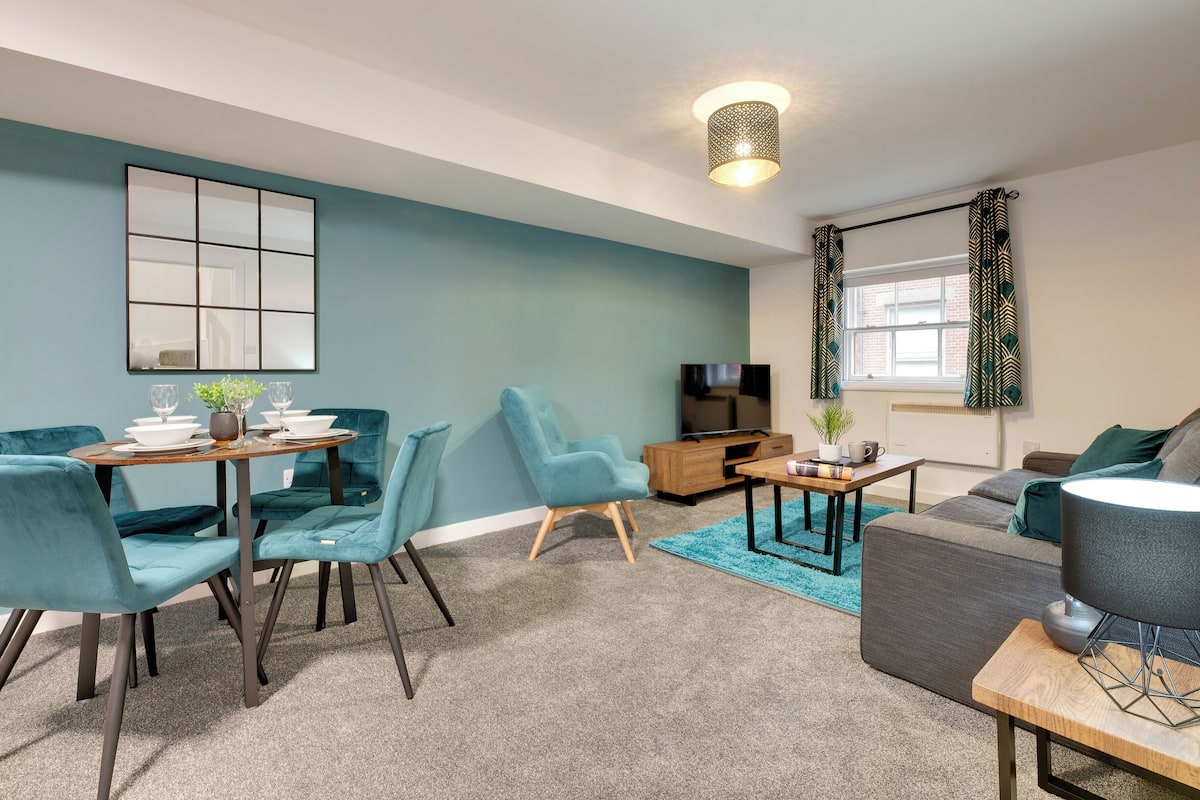 A modern living room with teal walls, a dining area with teal chairs, a grey sofa, and a wooden coffee table.