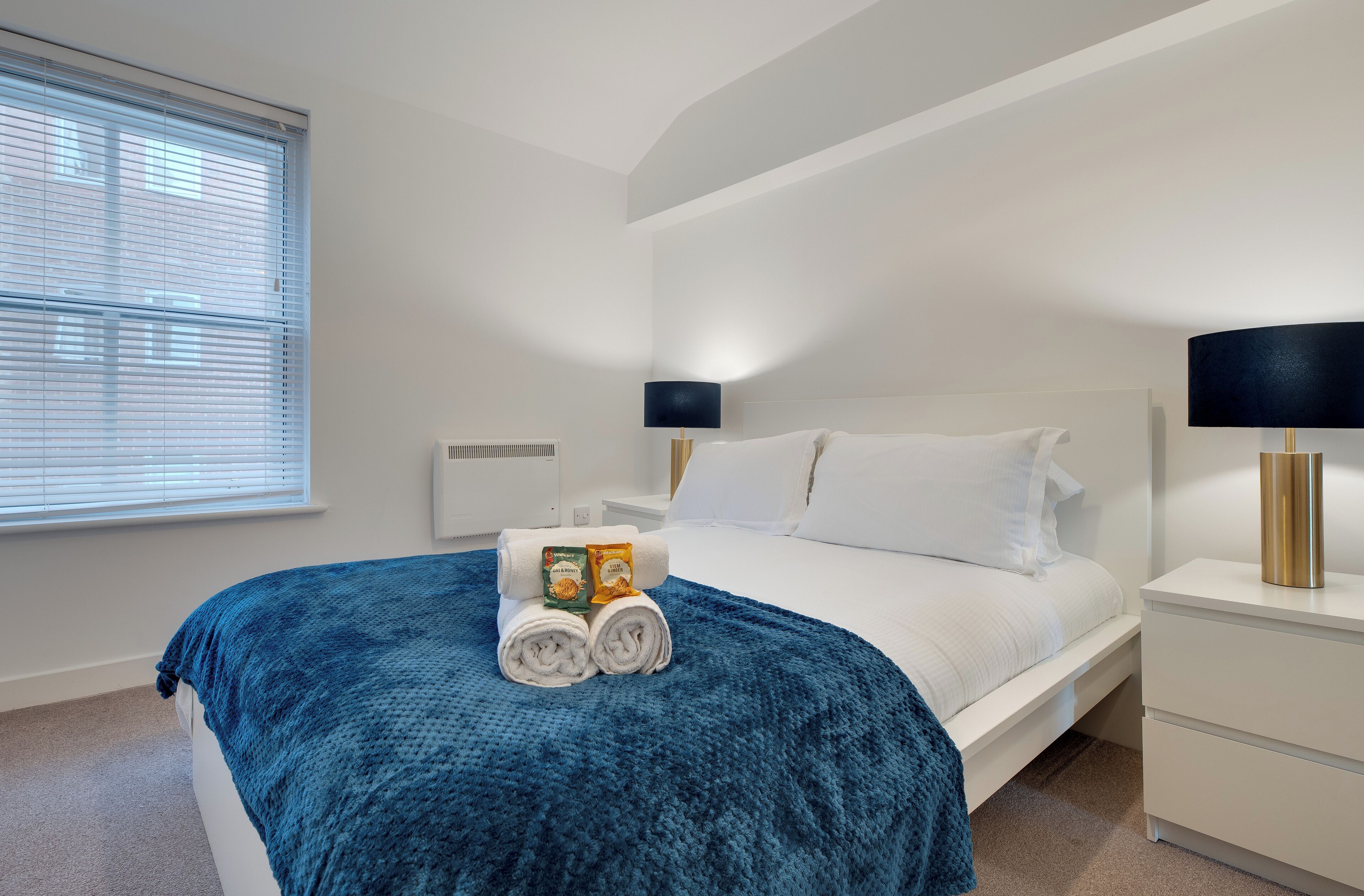 A neatly made bed with a blue blanket, a bedside lamp, and a rolled towel on the nightstand.