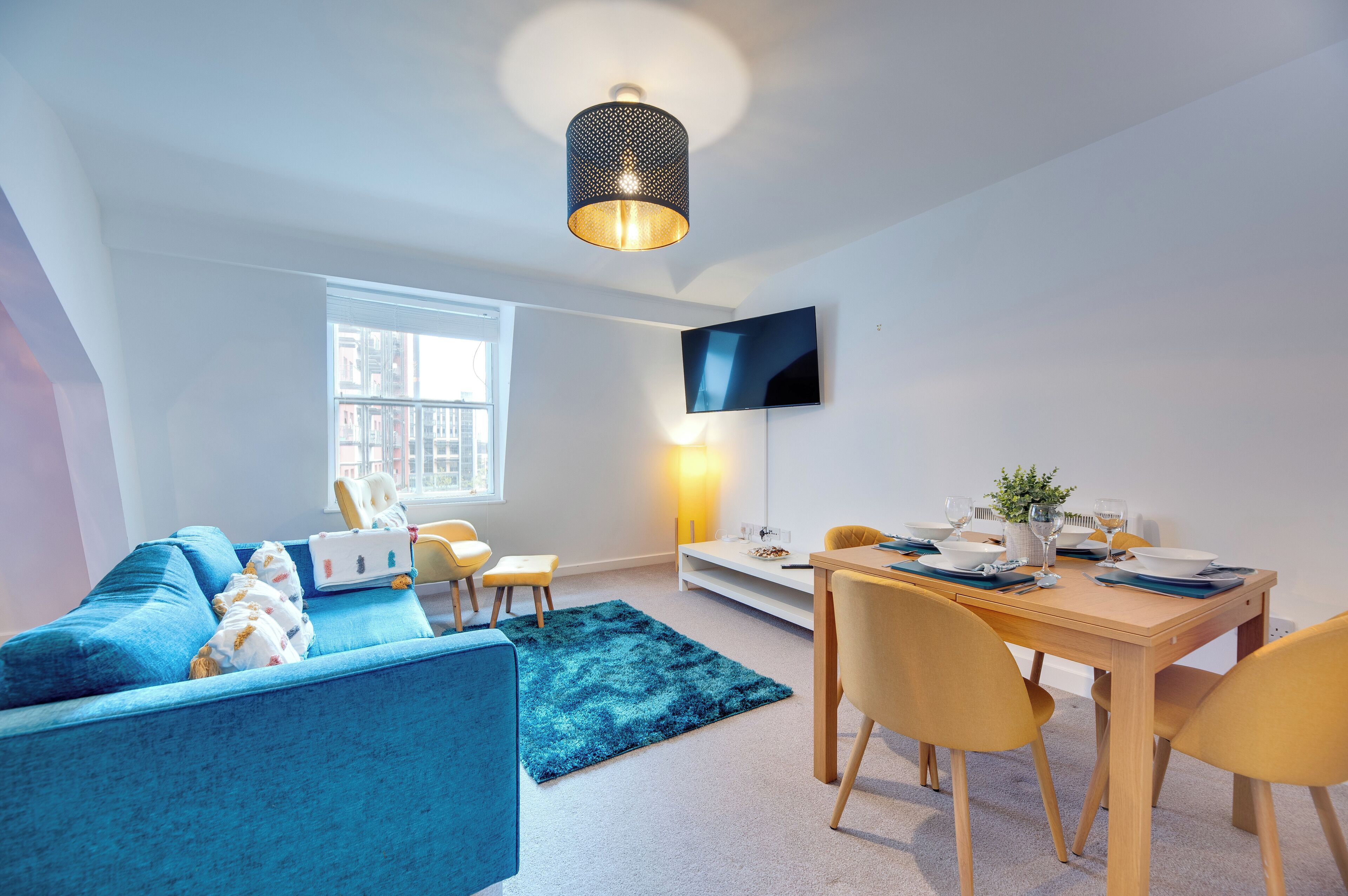 A modern living room with a blue sofa, a dining table with yellow chairs, a TV mounted on the wall, and a large window providing natural light.