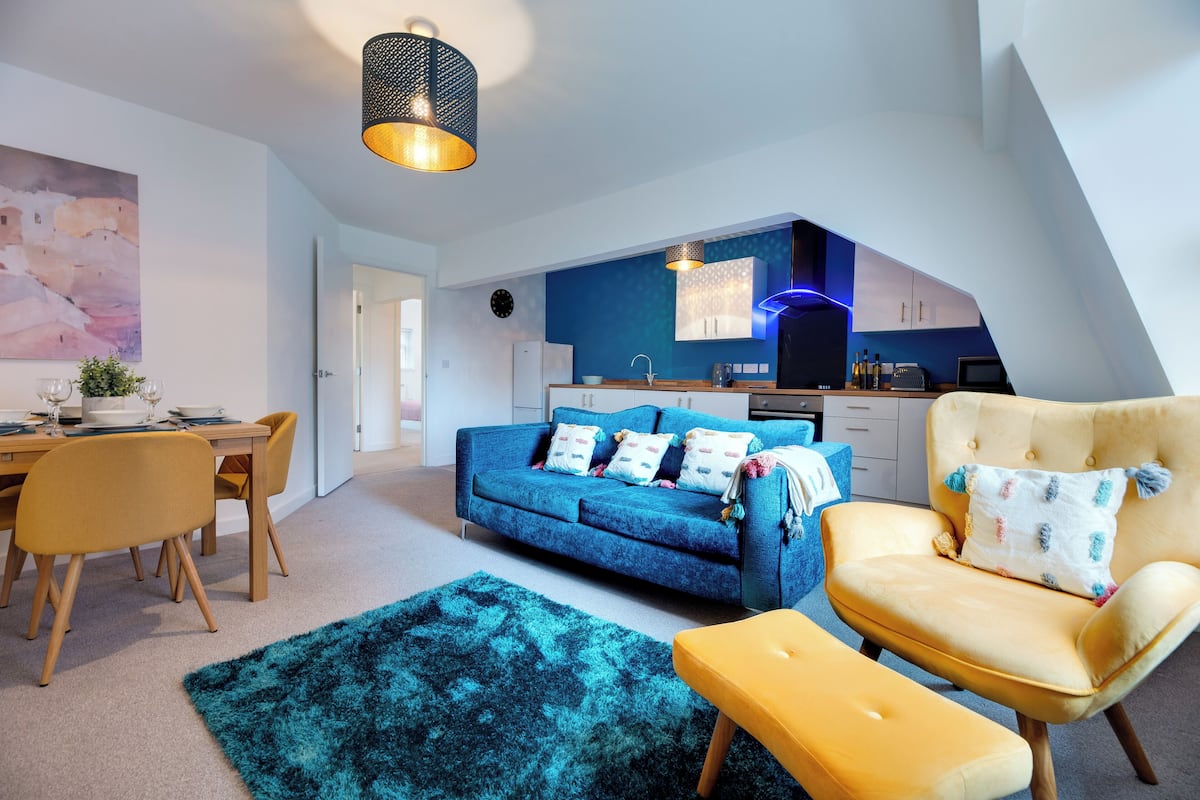 A modern living room with a blue sofa, yellow armchair, and a dining area with wooden chairs and a table.