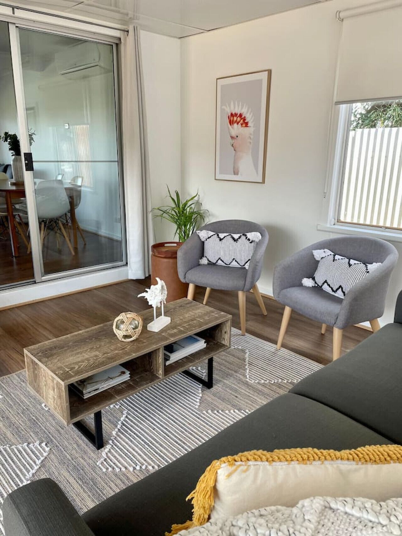 A modern living room with a large sliding glass door, a grey sofa, two armchairs, a wooden coffee table, and a framed artwork on the wall.