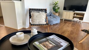 Un salon avec une table basse noire, un magazine, une bougie blanche et un fauteuil avec un coussin.