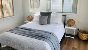 Une chambre à coucher avec un lit, des tables de chevet blanches, des paniers en osier et une fenêtre avec des stores.