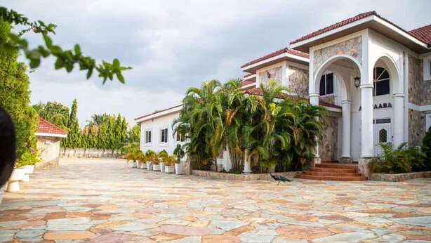 A spacious courtyard with a stone floor, surrounded by palm trees and a building with a tiled roof. - The Haven Boutique Hotel - Kumasi, Ghana (Ejisu)