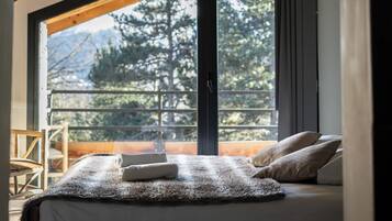 A bedroom with a large window, wooden beams, a bed with a fluffy blanket, and a view of trees and mountains.