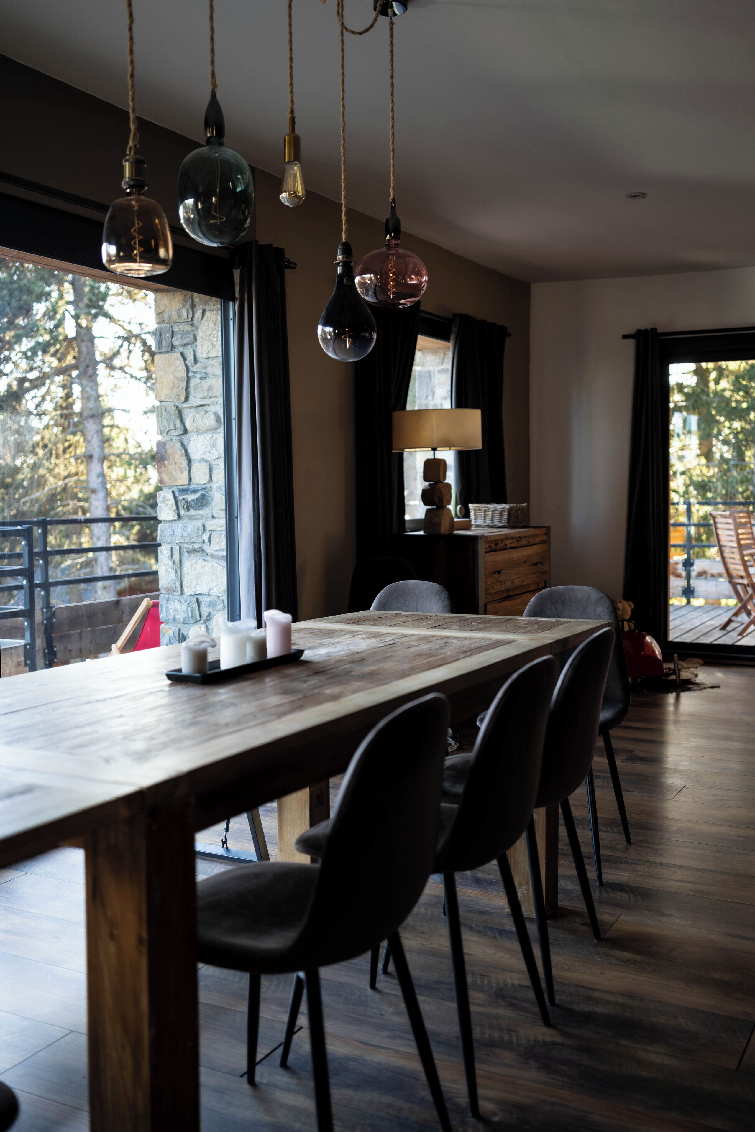 A dining area with a wooden table, black chairs, hanging pendant lights, and a view through a large window.